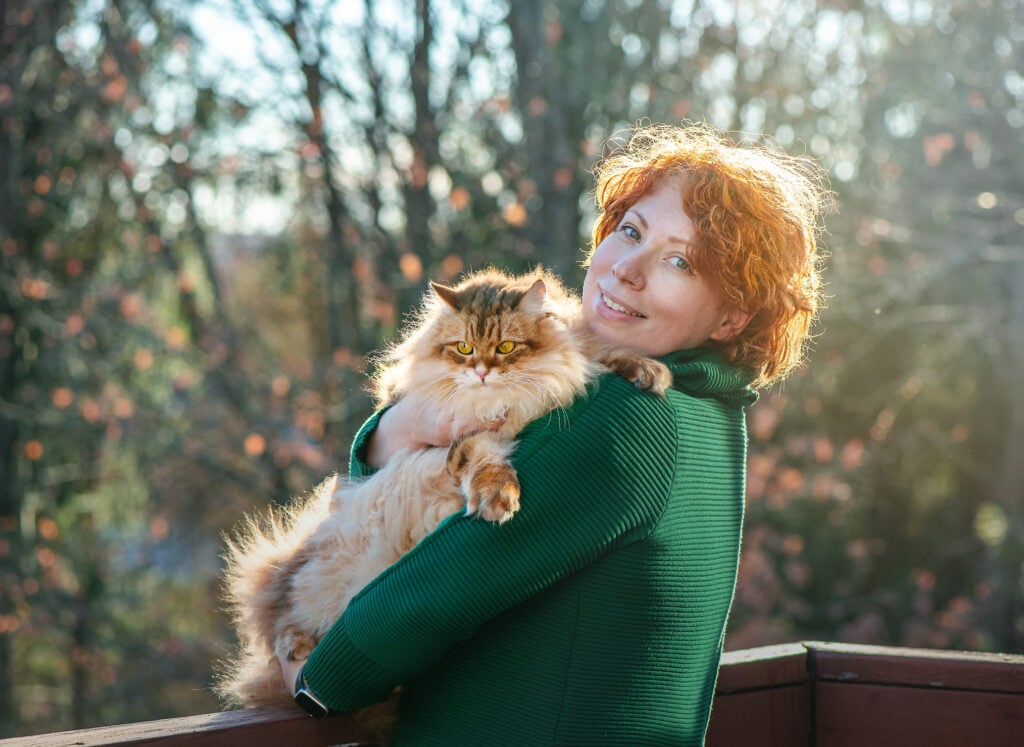 Woman with vibrant red hair cradles a long-haired Siberian cat in her arms on an outdoor deck. She wears a green sweater and smiles warmly while holding the fluffy feline. Sunlight filters through trees in the background, creating a soft, natural ambiance. Scene captures a moment of companionship between human and pet in a serene outdoor setting.
